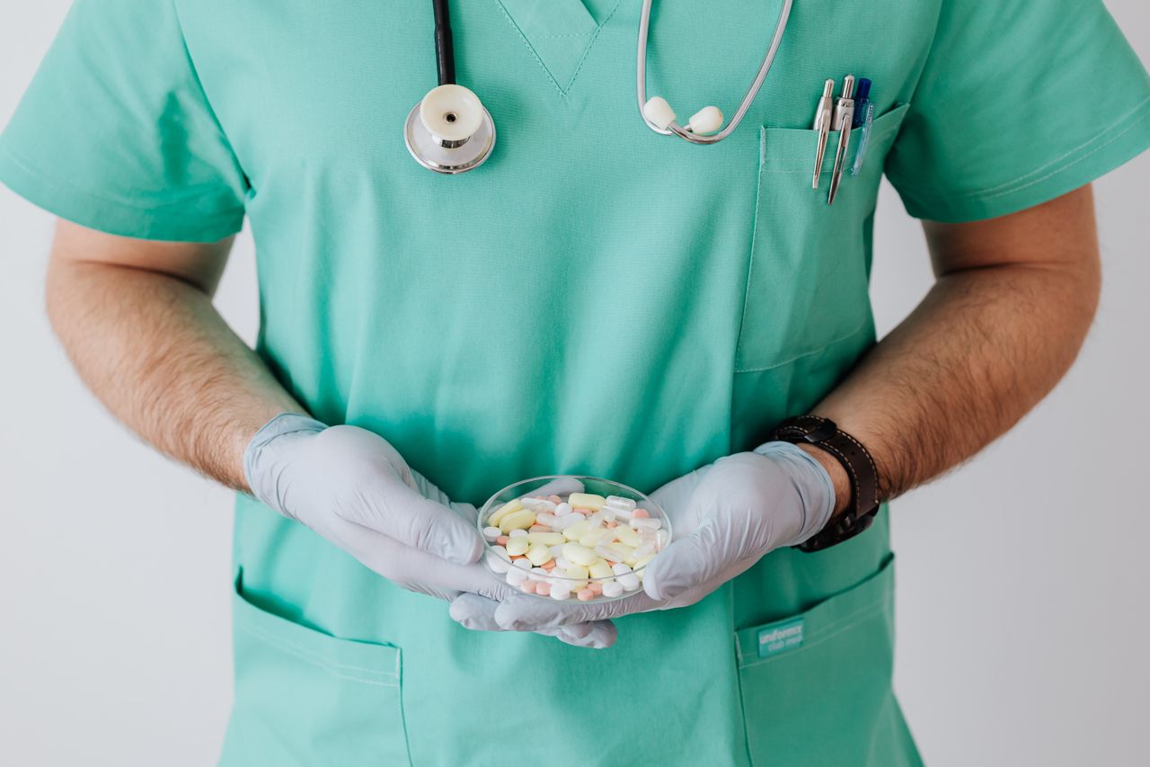 services-03 Unrecognizable male doctor wearing medical uniform and latex gloves holding Petri dish filled with assorted drugs while standing near white wall in hospital