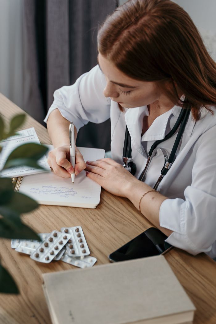 about-us-01 Young female doctor writing a prescription in a medical office with a stethoscope and pills nearby.